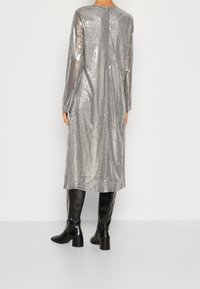 Robe en sequins argentés avec des manches longues, une coupe droite et une fermeture au dos, associée à des bottes en cuir noires hautes jusqu'aux genoux. Texture lisse.