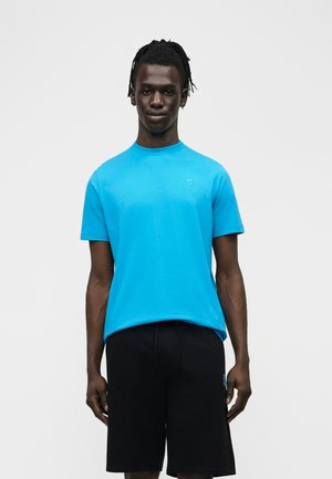 Man with braided hair wearing a bright blue short-sleeve T-shirt and black shorts standing against a plain light background.