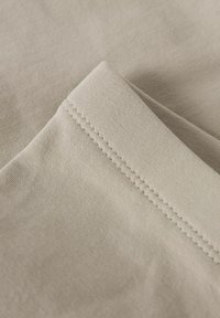 Beige stof met fijne textuur, met een close-up van een naad en een gevouwen rand, waarin de stikdetails en het gladde oppervlak worden benadrukt.