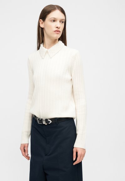 Femme aux longs cheveux bruns portant un pull blanc côtelé avec un col orné de perles et un pantalon noir ceinturé, regardant sur le côté.