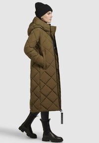 Long olive green quilted puffer coat with a high collar, zip closure, side pockets, and a slight sheen; paired with black knee-high boots.