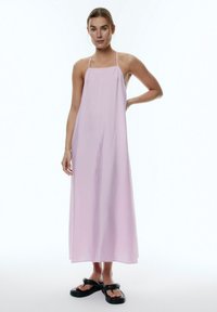 Helles rosa Maxikleid mit dünnen Trägern, das eine einfache A-Linie-Silhouette und strukturierten Stoff aufweist. Kombiniert mit schwarzen Plateausandalen.