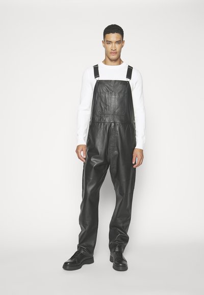 Zwarte leren tuinbroek met verstelbare banden, brede pijpen en een detail van een voorzak. Gecombineerd met een witte lange mouw shirt en zwarte schoenen.