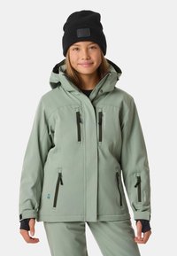 Jasnozielona, wodoodporna kurtka narciarska z kapturem, zamykanymi kieszeniami i regulowanymi mankietami. Nosi się z czarną czapką beanie.