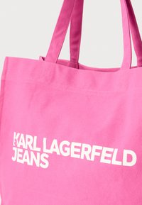 Ροζ τσάντα ώμου από καμβά με δύο λαβές, με εμφανές λευκό κείμενο "KARL LAGERFELD JEANS" στη μία πλευρά. Ανθεκτικός και απλός σχεδιασμός.