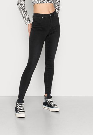 Jeans negros ajustados con cintura alta, que presentan una textura suave y un diseño estándar de cinco bolsillos. Combinados con zapatillas altas de color azul marino.