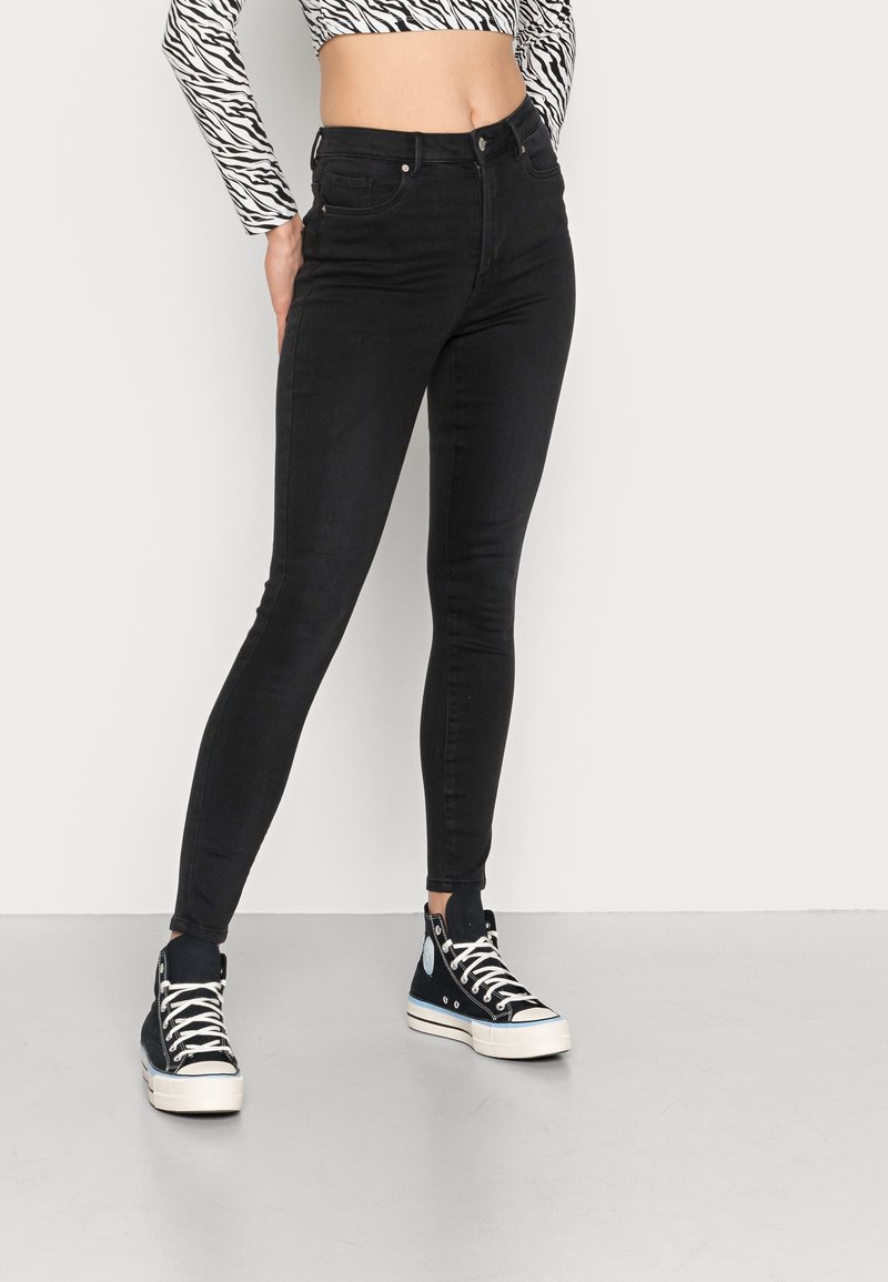 Jeans negros ajustados con cintura alta, que presentan una textura suave y un diseño estándar de cinco bolsillos. Combinados con zapatillas altas de color azul marino.
