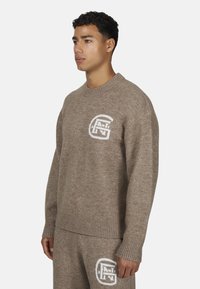 Młody mężczyzna w brązowym swetrze z dzianiny i spodniach z dopasowanymi białymi inicjałami logo na piersi i udzie, stojący na jednolitym tle.