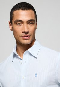 Ljusblå randig skjorta med en krispig krage, prydd med vita knappar och en liten blå logotyp. Slät tygtextur, skräddarsydd passform.