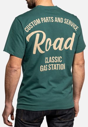 Homme portant un t-shirt vert avec un texte beige : « Custom parts and service Road Classic Gas Station » dans le dos.
