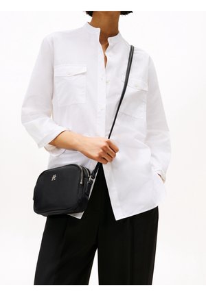 White, long-sleeve button-up shirt with two chest pockets, paired with a small black crossbody bag featuring a zipper and logo detail.