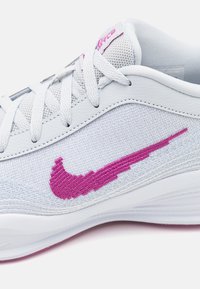 Bílé sportovní tenisky s textilním svrškem a texturovanými detaily. Mají výrazné růžové logo Nike, bílé tkaničky a polstrovaný jazyk.