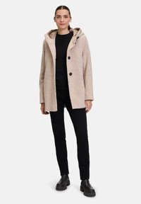Beige, fusseliger Mantel mit Kapuze, der schwarze Knöpfe und eine leicht übergroße Passform aufweist. Trägt einen schwarzen Oberteil und kombiniert mit schwarzen Hosen.