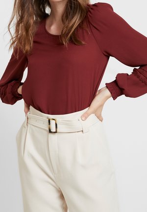 Femme portant un chemisier bordeaux à manches longues bouffantes et un pantalon crème taille haute avec une ceinture assortie.