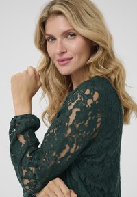 Top in pizzo verde scuro con motivi floreali e maniche trasparenti, caratterizzato da un colletto rotondo e dettagli di polsini arricciati.