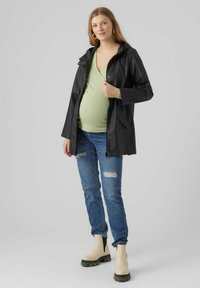 Zwarte regenjack met capuchon, sluiting met drukknopen aan de voorkant en zijzakken, gedragen over een lichtgroene top. Blauwe distressed jeans en beige enkellaarsjes.