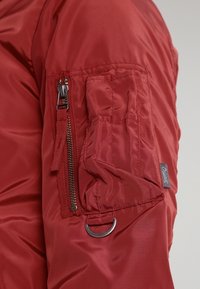 Czerwona nylonowa kurtka z zamkiem błyskawicznym na rękawie, akcentem w postaci pierścionka i metką. Materiał ma gładką, błyszczącą teksturę.