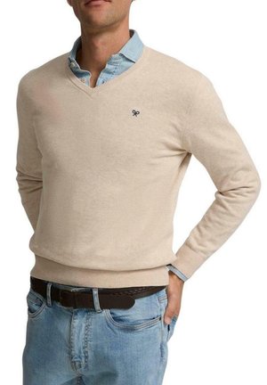 Suéter beige de pico en un material de punto suave, con un pequeño logo oscuro en el pecho, combinado con una camisa de cuello azul claro debajo.
