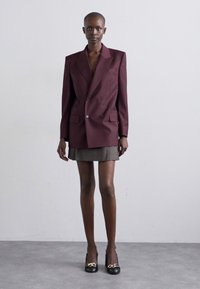Burgundy double-breasted blazer with sharp lapels, paired with a gray mini skirt. Model wears black loafers with gold hardware accents.