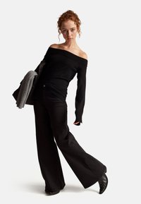 Černý top bez ramínek s dlouhými rukávy ve spojení s černými rozšířenými kalhotami. Textura vypadá hladce; modelka drží šedý šál.