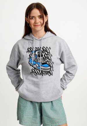 Jonge vrouw draagt een lichtgrijze hoodie met een blauwe sneakerafbeelding en groen-wit gestreepte shorts, glimlachend met handen in de zakken.