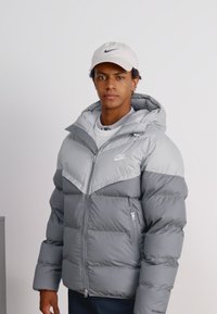 Casaco de penas cinza com um design em forma de zigue-zague, equipado com capuz, bolsos com fecho e um logótipo. Textura suave e acabamento ligeiramente brilhante.