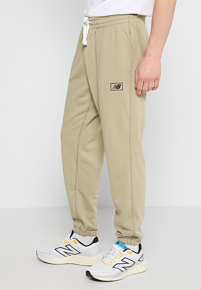 Pantalons de survêtement beiges avec une taille à cordon de serrage et des détails de couture sur les côtés. Patch logo sur la cuisse. Portés avec des chaussures de sport blanches et jaunes.