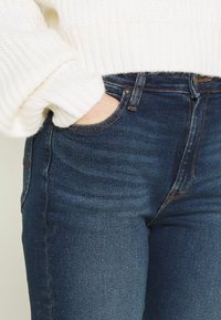 Fitted dark blue denim jeans with a smooth texture, featuring minimal fading, functional pockets, and contrasting orange stitching details.