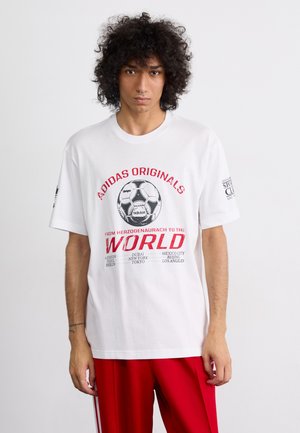 Hvid bomuld t-shirt med et stort sort og rødt fodboldgrafik og tekst, kortærmet og afslappet pasform. Modellen har røde bukser på.