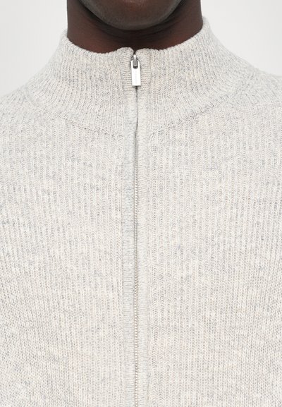 Ljusgrå stickad tröja med hög krage, som har en dragkedja framtill. Texturen är ribbad och visar ett subtilt mönster.