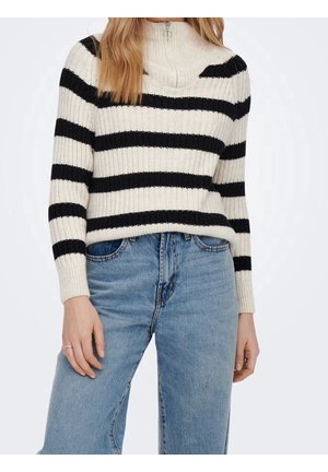 Femme portant un pull côtelé blanc avec des rayures horizontales noires et un col zippé, associé à un jean bleu taille haute.