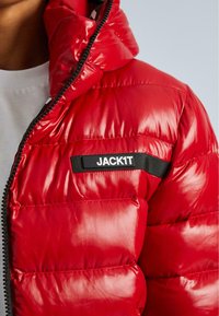 Közelkép egy személyről, aki fényes piros steppelt kabátot visel fekete zipzárral és egy "JACK1T" címkével a mellkasán.