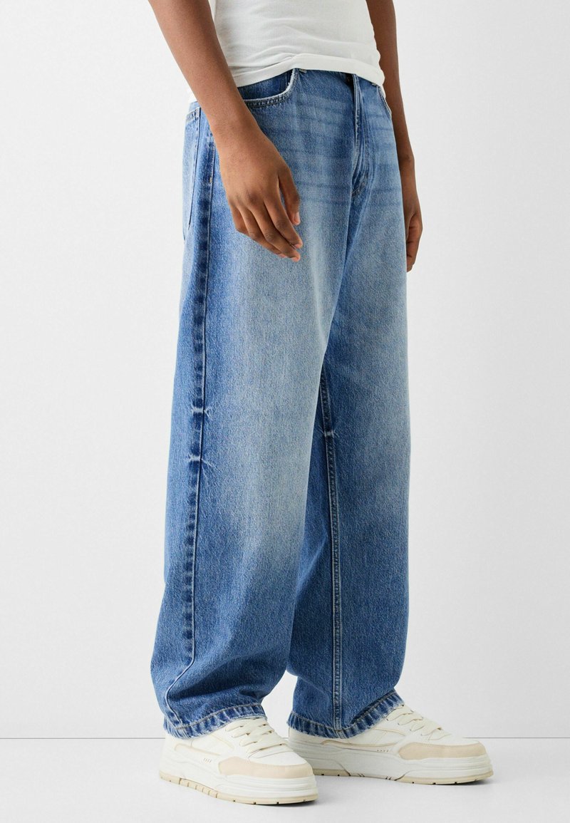 Jeans a gamba larga in denim blu con una vestibilità rilassata, caratterizzati da una finitura sbiadita, dettagli strappati e il classico stile a cinque tasche. Abbinati a sneakers bianche.