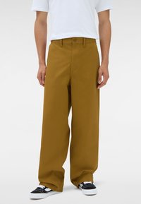 Uomo che indossa pantaloni larghi senape, una maglietta bianca e sneaker bianco e nero, in piedi contro uno sfondo bianco uniforme.
