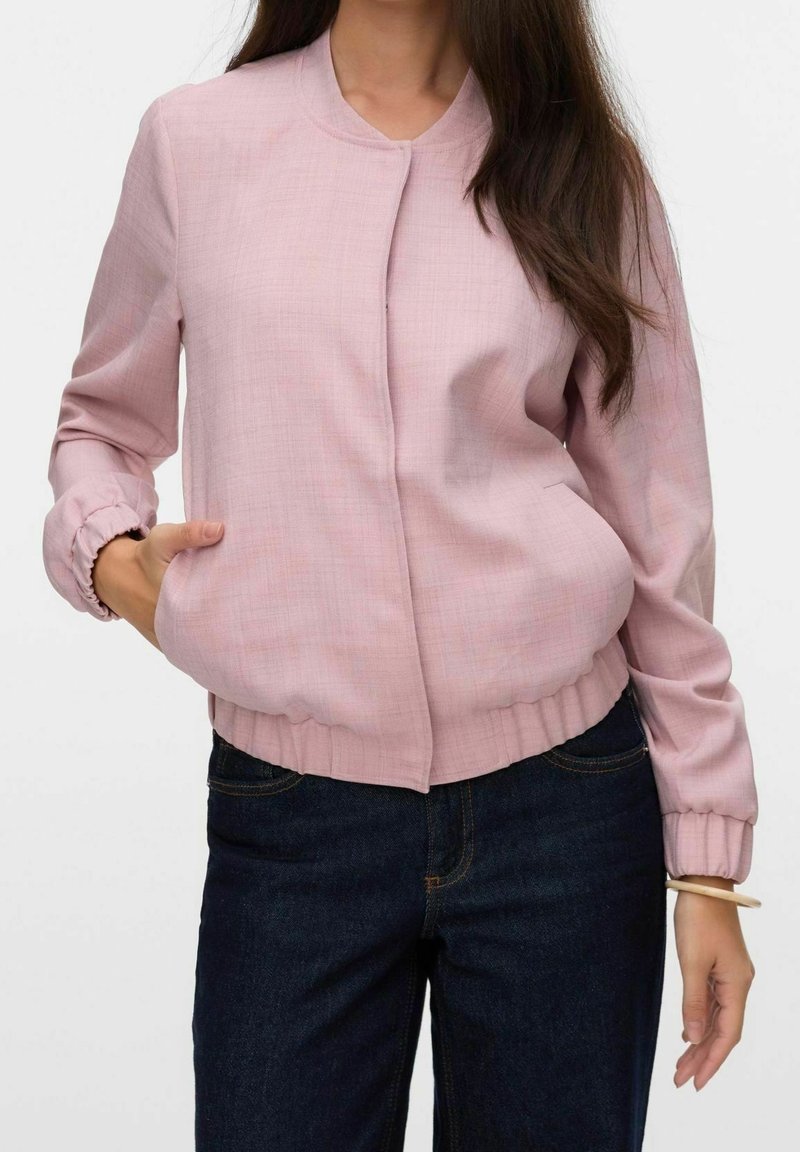 Femme portant une veste bomber rose clair avec poignets et ceinture élastiques, associée à un jean bleu foncé, posant avec une main dans la poche.