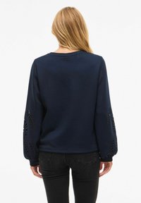 Donna con lunghi capelli biondi che indossa una felpa blu navy con ritagli floreali sulle maniche e jeans neri, vista di spalle.