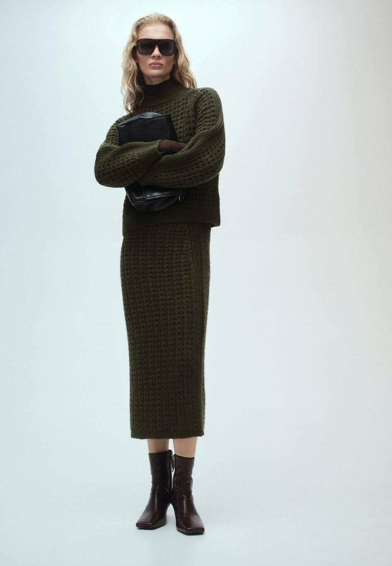 Dunkelgrünes gestricktes Oberteil und Midi-Rock-Set mit strukturiertem Muster; kombiniert mit schwarzer Handtasche und braunen Stiefeletten. Modell trägt eine Sonnenbrille.