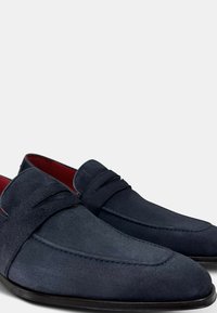 Mocasines de ante azul marino con detalles cosidos y un acabado suave; forro interior rojo; suela negra de bajo perfil; diseño clásico.