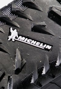 Svart gummidäck med framträdande, vinklade sulor för grepp, med en tydlig Michelin-logotyp och en texturerad yta.