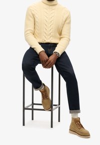 Pull à col roulé beige en maille torsadée, jean en denim foncé, et bottes en daim marron clair avec des lacets bruns. Le pull présente des motifs texturés et des poignets côtes.