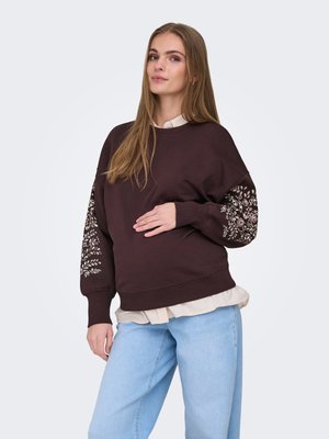 Giovane donna con i capelli lunghi che indossa una felpa marrone scuro con motivi floreali sulle maniche, una camicia beige chiaro e jeans azzurri.