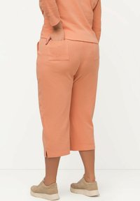 Ferskenfarvede tætsiddende bukser med to baglommer, sideslidser ved kanten, lavet af blødt materiale. Båret med lysebeige sneakers.