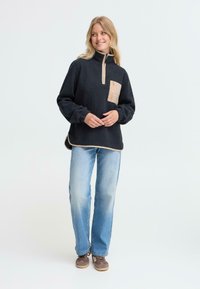 Svart fleecepullover med halvlending og krage, beige brystlomme og sidesplitt, parret med lyseblå jeans og brune sko.
