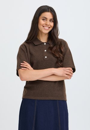 Jeune femme aux longs cheveux foncés portant un haut marron à manches courtes avec trois boutons et une jupe bleue, souriante les bras croisés.