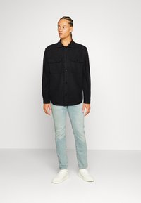 Sort skjorte med knapper og to brystlommer, let stof, tætsiddende design, parret med lyseblå jeans og hvide sneakers.