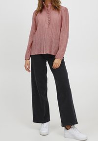 Rosa, strukturierte Bluse mit Knopfleiste und Rüschendetail, kombiniert mit schwarzen weiten Hosen und weißen Turnschuhen.