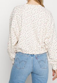 Krémová košile s dlouhými rukávy a malým růžovým květinovým vzorem, s elastickým pasem, spárovaná se světle modrými džíny s vysokým pasem a zadními kapsami.