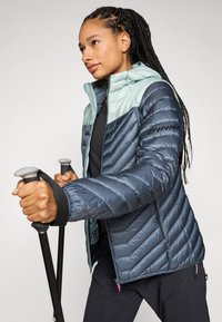 Jachetă căptușită de culoare albastră închisă cu accente de albastru deschis. Are un fermoar frontal, glugă și o textură matlasată în diagonală. Logo negru pe mânecă.