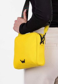 Žlutá plátěná taška přes rameno s černým logem, která má strukturovaný design a vícebarevný vzorovaný popruh. Zapínání na zip, obdélníkový tvar.