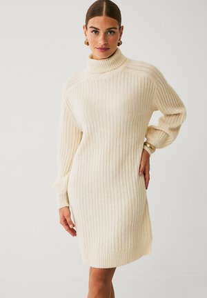 Gebreide jurk in crème met ribbeltextuur, hoge col, relaxte pasvorm en lange mouwen. Zijsplitten aan de zoom voor een gemakkelijke beweging.